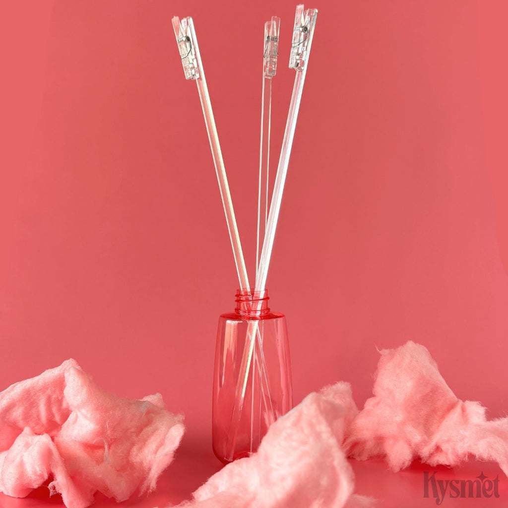 Three plastic sticks with clear clothespins attached are sticking out of a pink bottle surrounded by pink cotton candy against a pink background.