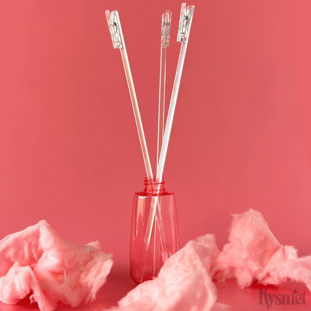 Three plastic sticks with clear clothespins attached are sticking out of a pink bottle surrounded by pink cotton candy against a pink background.