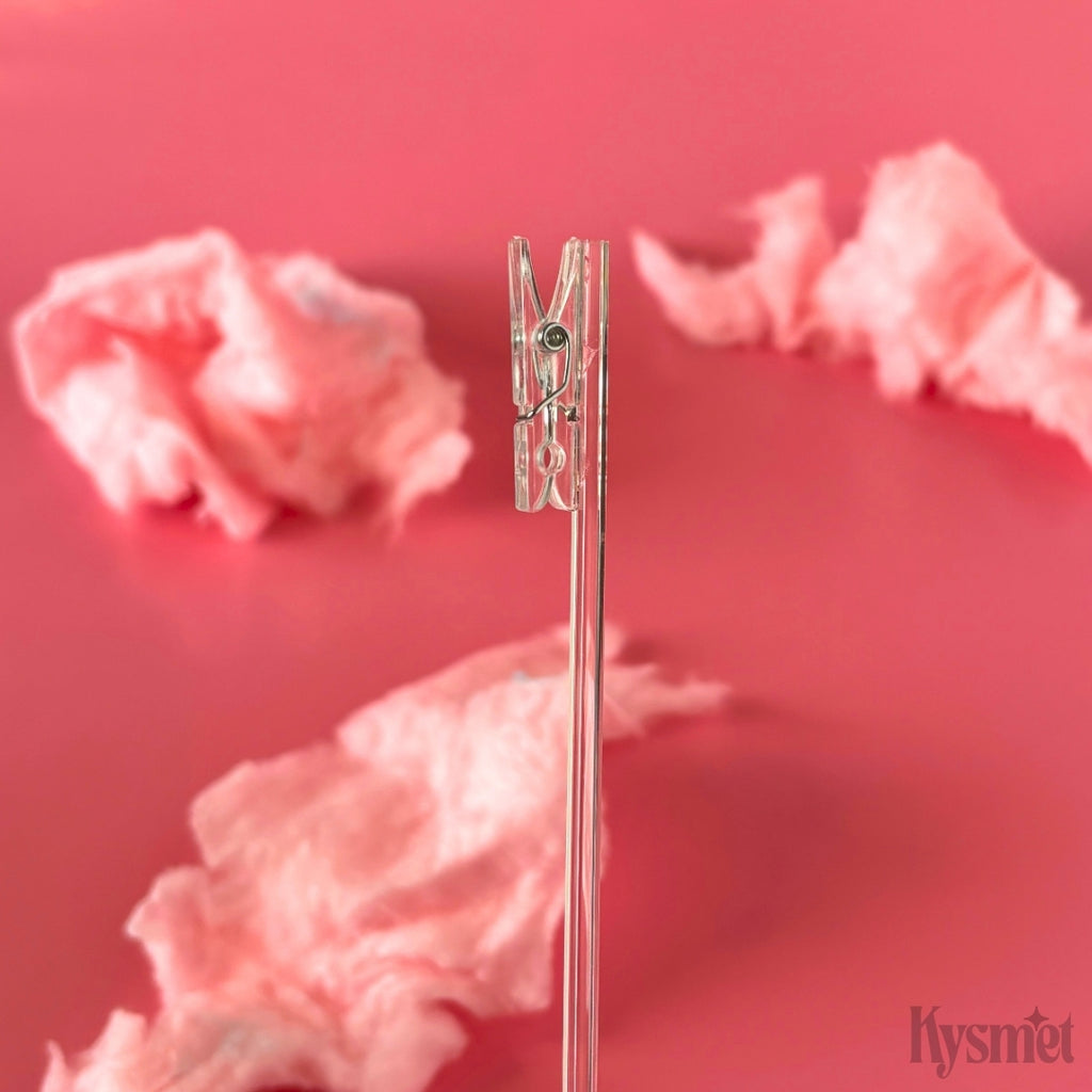 A clear plastic stick with a clear clothespin is seen against pink cotton candy and a pink background.
