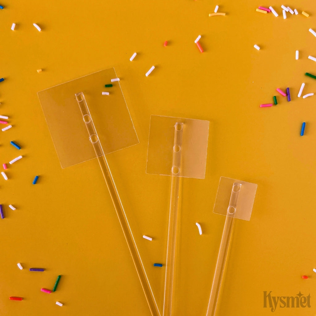 Three plastic sticks with clear squares attached are surrounded by rainbow sprinkles against an orange background.